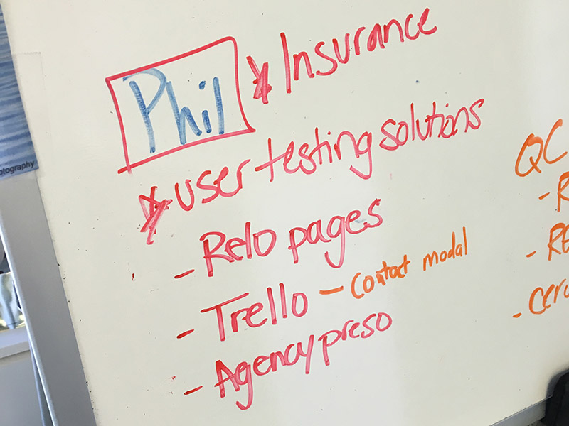 A list of tasks written on a whiteboard, reading: Phil- Insurance, User testing solutions, Relo pages, Trello, agency preso